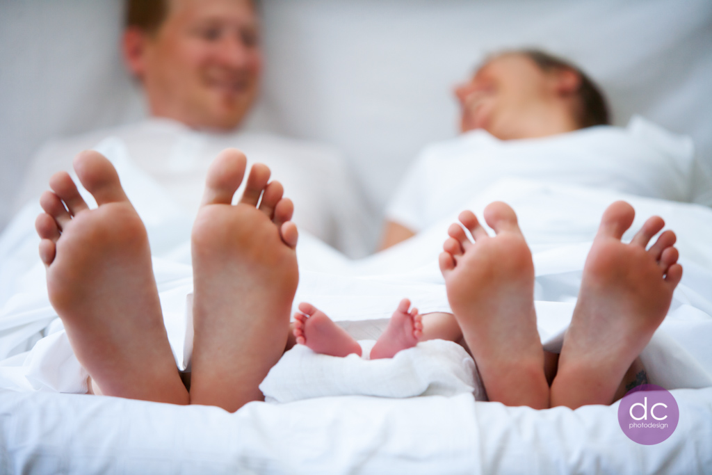 Neugeborenes Fotoshooting bei dem Eltern mit ihrem Baby im Bett liegen und ihre Füße unter der Bettdecke zeigen