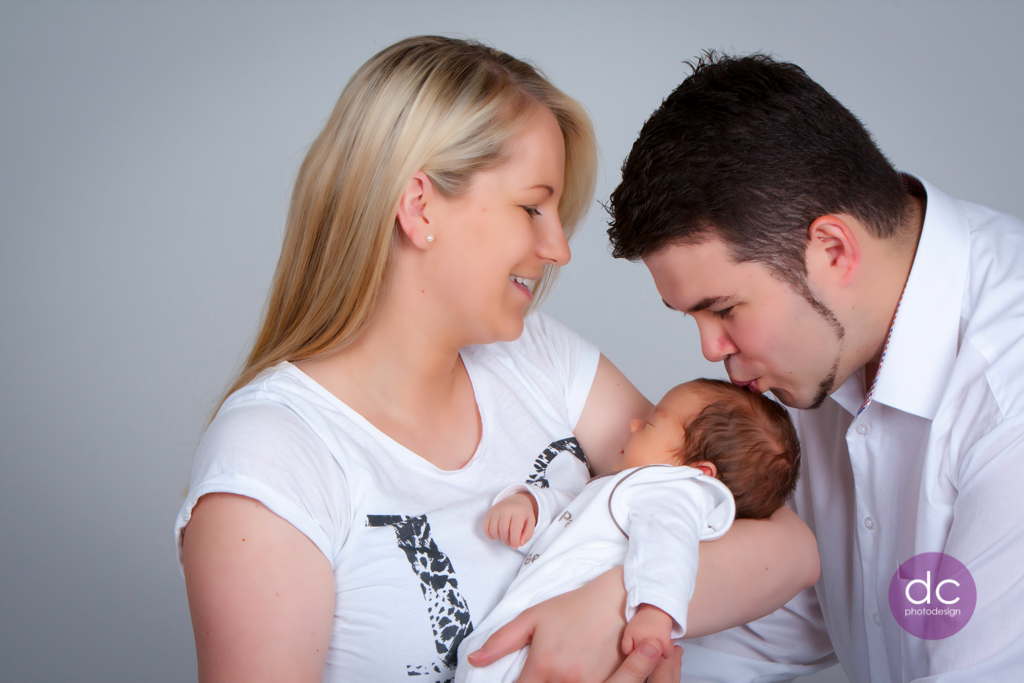 Neugeborenes Fotoshooting Mutter mit ihrem Baby auf dem Arm und Papa gibt einen Kuss auf den Kopf
